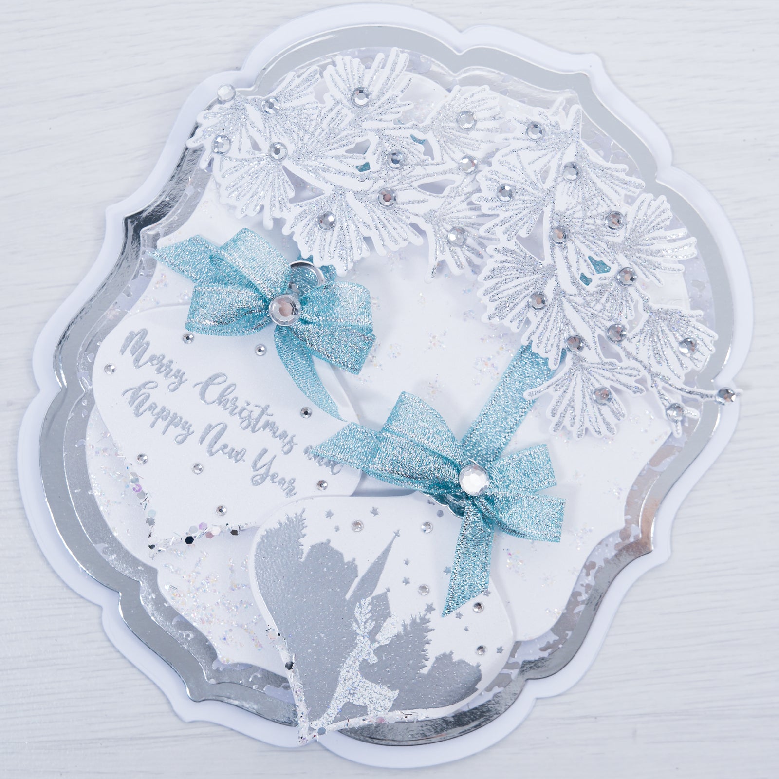 Learn how to make this fancy easel style Christmas card. This beautiful blue and silver toned card features crystal encrusted foliage and snow scene baubles with luxe ribbon bows.